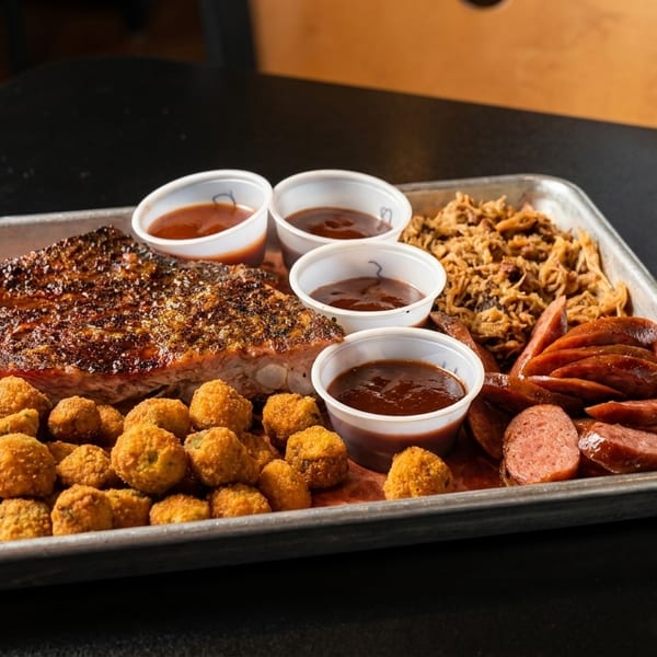 A metal tray with BBQ ribs, fried okra, pulled pork, sliced sausage, and four cups of sauce.